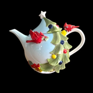May include: A ceramic Christmas tree teapot with a light blue body and a white handle and spout. The teapot is decorated with a green Christmas tree, red cardinals, and colorful ornaments. A white star sits atop the tree.