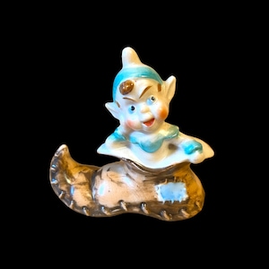 May include: A ceramic figurine of an elf wearing a blue and white hat and sitting in a brown boot. The elf has a smiling face and brown eyes.