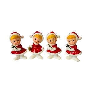 May include: Set of four vintage ceramic Christmas figurines. Each figurine depicts a girl wearing a red and white Santa suit holding a gift.