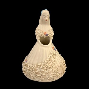 May include: A white ceramic figurine of a woman in a lace dress. The dress has a floral design and a small opening in the front for holding flowers.
