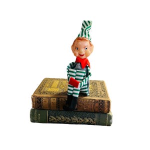 May include: A vintage Christmas elf doll with a red collar, green and white striped hat and outfit, and black boots. The elf is sitting on top of two antique books with gold lettering and green covers.