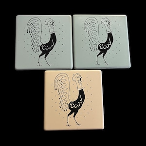 以下が含まれることがあります： 正方形のセラミックコースター3枚。2つは水色で、1つはクリーム色です。各コースターには、大きな尾を持つ雄鶏の黒いイラストと、小さな黒い十字の装飾的なパターンが描かれています。