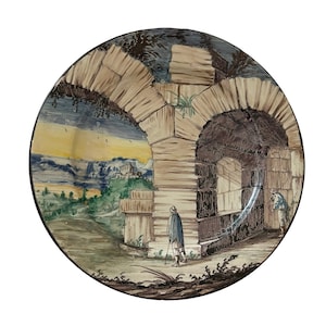 May include: A ceramic plate featuring a hand-painted scene of a stone archway with two figures walking through it. The scene is set in a landscape with mountains in the background. The plate is decorated in a yellow, blue, and green colour scheme.