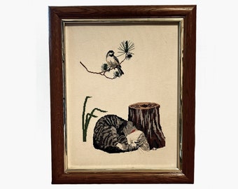 May include: A framed cross-stitch embroidery depicting a sleeping cat curled up next to a tree stump. A small bird perches on a branch above the cat.