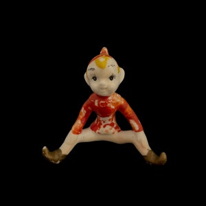 May include: A ceramic elf figurine with a red and white patterned outfit, sitting with legs spread wide and wearing brown shoes.