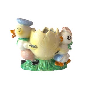 May include: A ceramic Easter planter featuring two duckling figures pushing a cracked eggshell. The ducklings wear sailor hats, scarves, and overalls. The planter is painted in pastel colours and sits on a patch of green grass.
