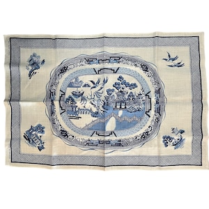 May include: A white tea towel with a blue and white willow pattern. The pattern features a bridge, trees, and a pagoda. The towel has a blue border.