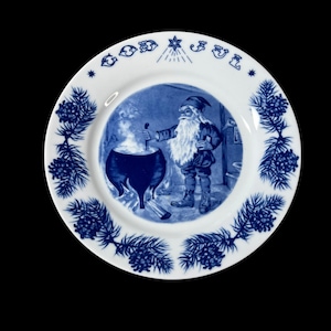 May include: A white and blue decorative plate featuring a blue illustration of a gnome stirring a cauldron. The plate has a blue pine cone and branch border, with the words "GOD JUL" in blue. The plate is round.