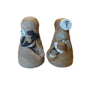 May include: Two ceramic salt and pepper shakers shaped like faces. The salt shaker has a mustache and a dollar sign on its eye. The pepper shaker has a gray beard and the letter "P" on its head.