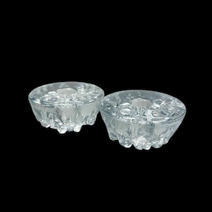 May include: Two clear glass candle holders with a textured, ice-like appearance. Each holder has a circular indentation for a candle and a rounded base with small, irregular bumps. The background is solid black.