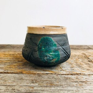 May include: A small, black ceramic cup with a beige rim and a teal-green glaze detail. The cup is sitting on a rustic wooden surface.