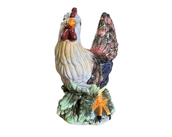 May include: A ceramic rooster figurine with a white, tan, and red body, and a colorful tail. The rooster stands on a base of green leaves. The beak is orange, and the comb is red.
