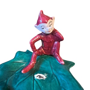May include: A ceramic elf figurine in a red outfit with a white face, seated on a large green leaf. The elf is resting its arm. The leaf has a glossy finish. A small label is visible on the leaf.
