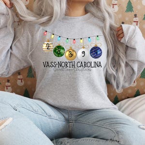May include: A gray sweatshirt with a Christmas ornament design. The ornaments have the numbers 2, 8, 3, 9, and 4 on them. The text on the sweatshirt reads "VASS-NORTH CAROLINA Small Town Christmas".