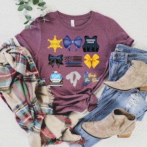 May include: A maroon t-shirt with a variety of blue and yellow police-themed graphics, including a police badge, a police vest, a police car, a blue ribbon, and a yellow ribbon. The text "Back the Blue" is also included on the shirt.