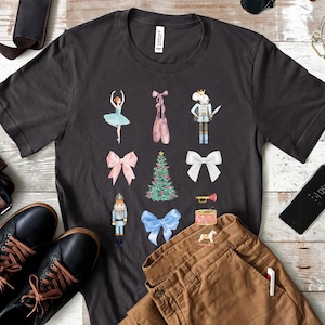 May include: A dark grey t-shirt with a Christmas Nutcracker theme. The design features a ballerina, a nutcracker, a Christmas tree, a toy rocking horse, a drum, a trumpet, and pink and blue bows.