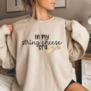 Könnte beinhalten: Beiges Sweatshirt mit dem Text "in my string cheese era" in schwarzer Schrift. Unter dem Text befindet sich eine Grafik von Fädenkäse, der mit einer rosa Schleife gebunden ist. Das Sweatshirt hat lange Ärmel.