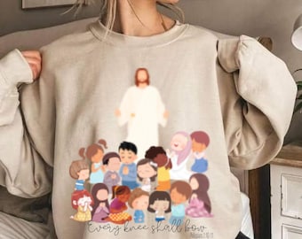 May include: A beige sweatshirt with a graphic print of Jesus surrounded by children. The text "Every knee shall bow Philippians 2:10-11" is printed below the graphic.