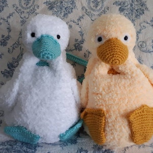 Könnte beinhalten: Zwei gehäkelte Plüsch-Entenspielzeuge. Eine Ente ist weiß mit einem hellblauen Schnabel und Füßen. Die andere Ente ist hellorange mit einem gelben Schnabel und Füßen.