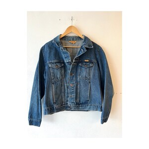May include: A medium-wash blue denim jacket featuring a classic collar, long sleeves, and a button-up front. The garment includes two chest pockets with buttoned flaps and a small rectangular label on the interior collar area reading Wrangler.