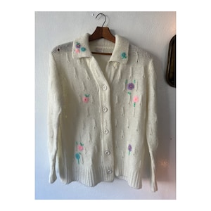 May include: A white knitted cardigan with a button-up front and a collar. The cardigan is decorated with embroidered flowers in shades of pink, purple, blue, and green.