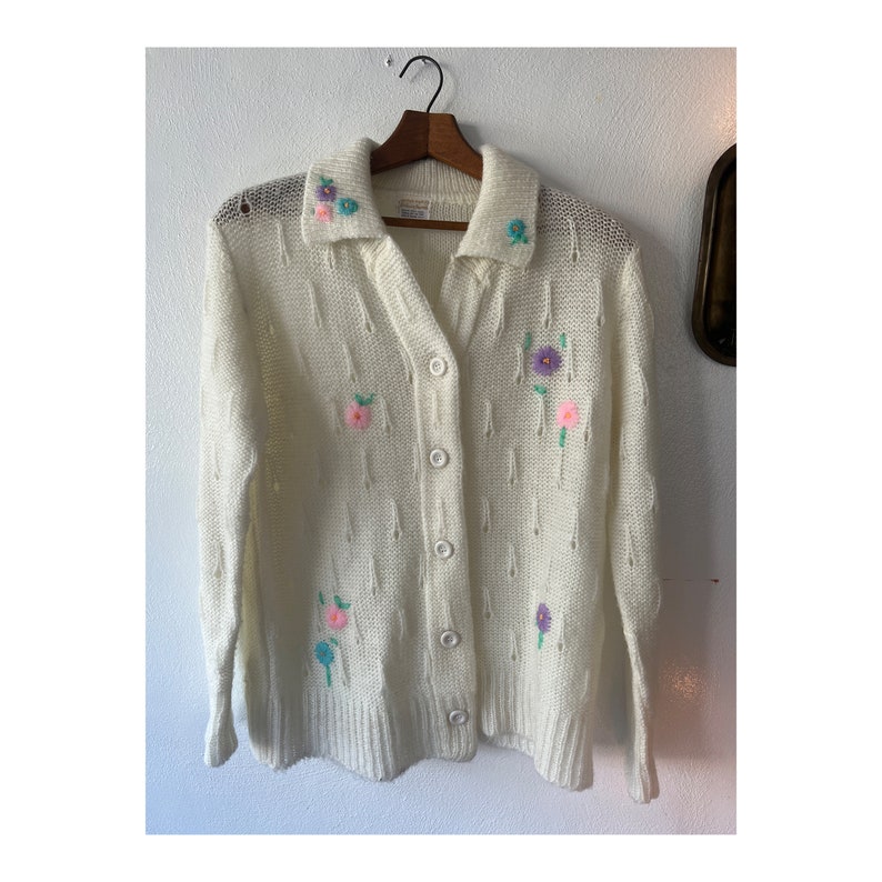May include: A white knitted cardigan with a button-up front and a collar. The cardigan is decorated with embroidered flowers in shades of pink, purple, blue, and green.