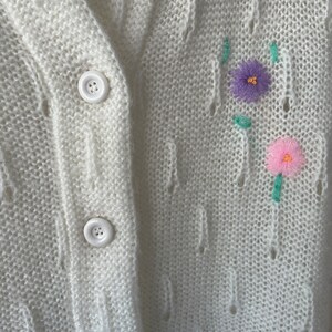 May include: White knitted cardigan with a cable knit pattern and embroidered flowers in pink, purple, and green.
