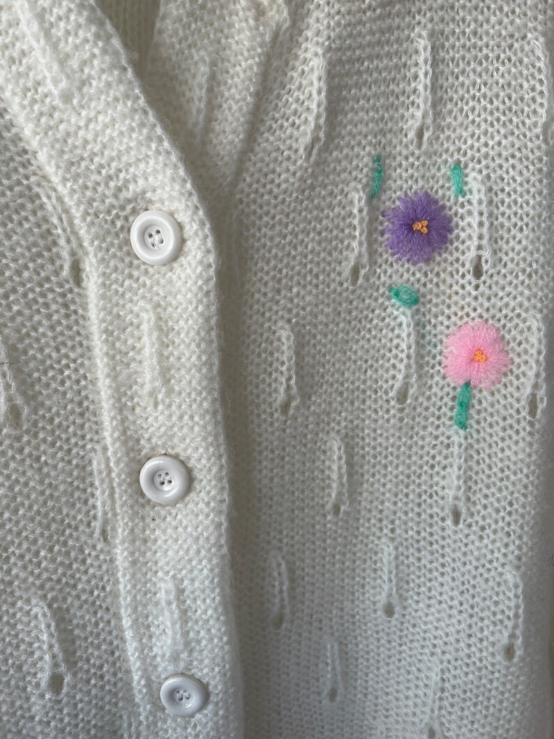 May include: White knitted cardigan with a cable knit pattern and embroidered flowers in pink, purple, and green.