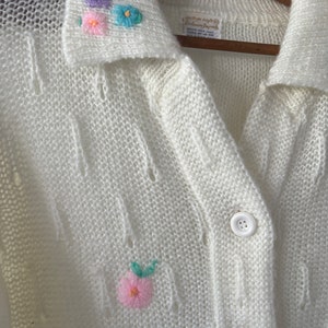 May include: White knitted cardigan with a collared neckline and embroidered flowers. The cardigan has a button closure and a delicate openwork pattern.