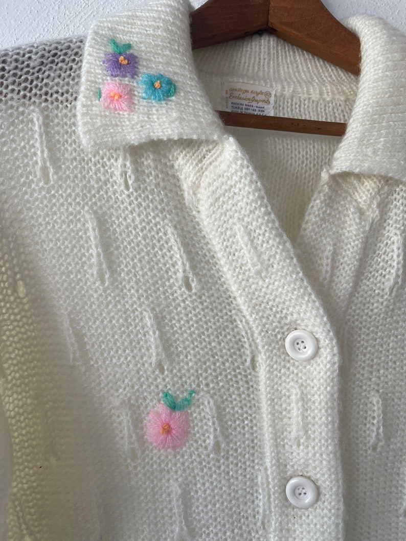 May include: White knitted cardigan with a collared neckline and embroidered flowers. The cardigan has a button closure and a delicate openwork pattern.