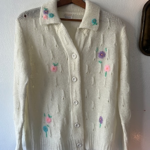 May include: A white knitted cardigan with a button-up front and a collar. The cardigan is decorated with embroidered flowers in shades of pink, purple, and blue.