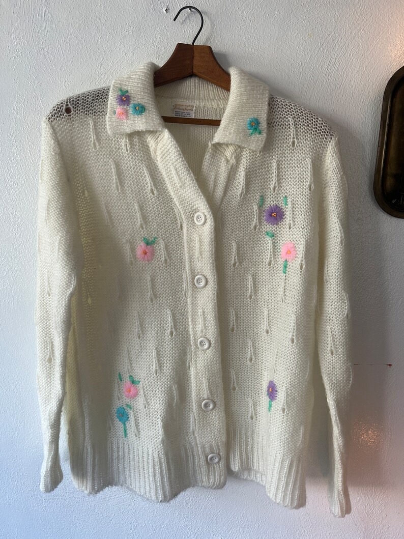 May include: A white knitted cardigan with a button-up front and a collar. The cardigan is decorated with embroidered flowers in shades of pink, purple, and blue.