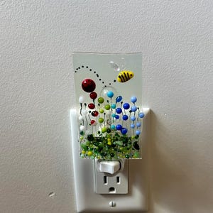 May include: A rectangular fused glass night light plugged into a wall socket. The glass features a clear background with a colourful garden scene of green, red, blue, and white glass beads and wire stems, topped with a small yellow and black bee.