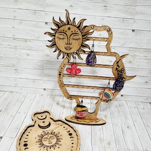 May include: Wooden earring stand with a sun and moon design. The stand has multiple slots for earrings and a small tray at the bottom. The stand is made of light brown wood and has a natural finish.