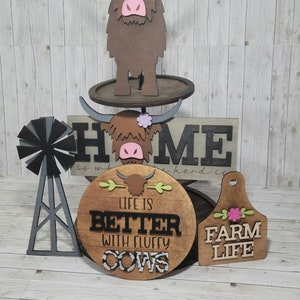 Op de afbeelding: Houten boerderijdecoratie met een hooglandkoe, een windmolen en drie borden. De borden lezen "HOME is where the herd is", "Life is better with fluffy cows" en "Farm Life".