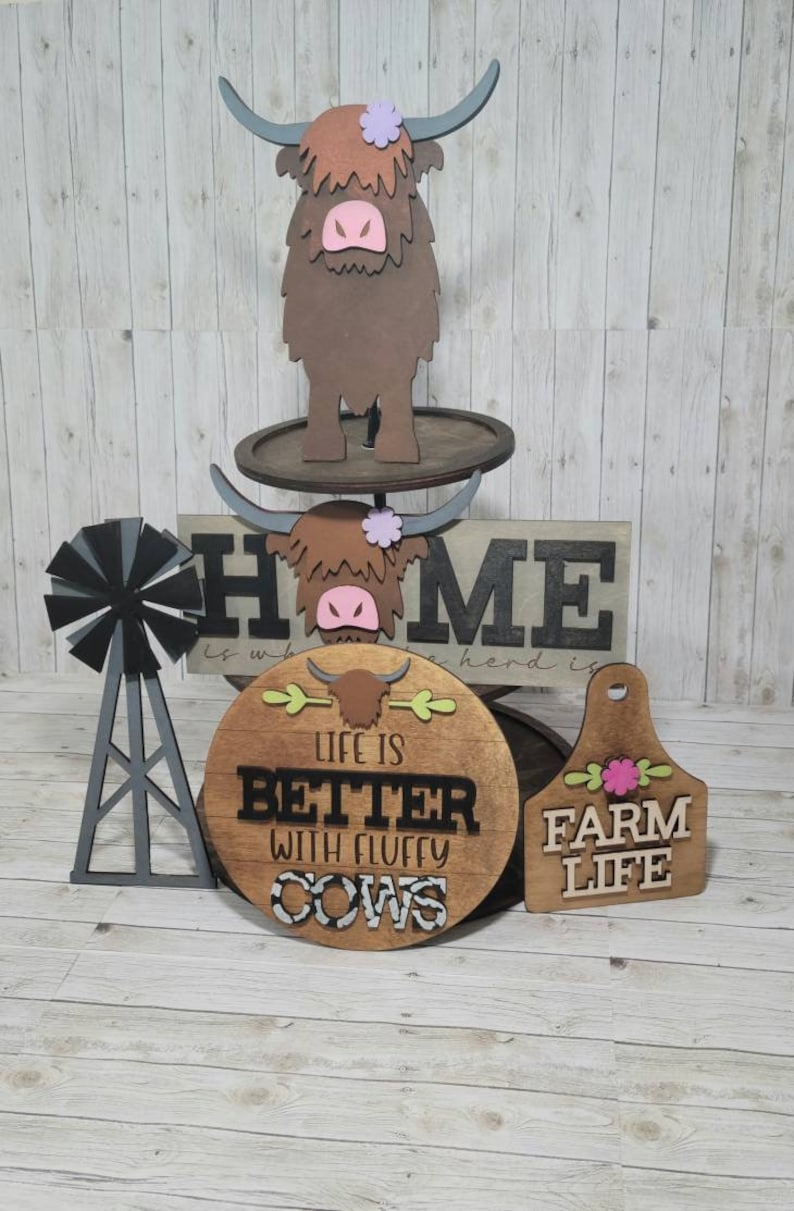 Op de afbeelding: Houten boerderijdecoratie met een hooglandkoe, een windmolen en drie borden. De borden lezen "HOME is where the herd is", "Life is better with fluffy cows" en "Farm Life".