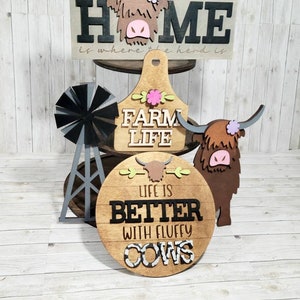 Op de afbeelding: Houten boerderijdecoratie met een zwart-wit bord met de tekst "Home is where the herd is", een bruin houten bord met de tekst "Farm Life", een bruin houten bord met de tekst "Life is better with fluffy cows", en een zwarte en grijze windmolen.