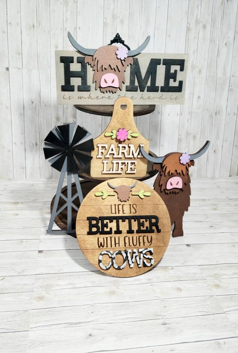 Op de afbeelding: Houten boerderijdecoratie met een zwart-wit bord met de tekst "Home is where the herd is", een bruin houten bord met de tekst "Farm Life", een bruin houten bord met de tekst "Life is better with fluffy cows", en een zwarte en grijze windmolen.