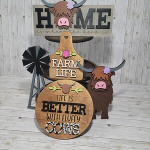 Op de afbeelding: Een set van drie houten borden met een boerderijthema. Het bovenste bord staat "Home is where the herd is". Het middelste bord staat "Farm Life" en het onderste bord staat "Life is better with fluffy cows".