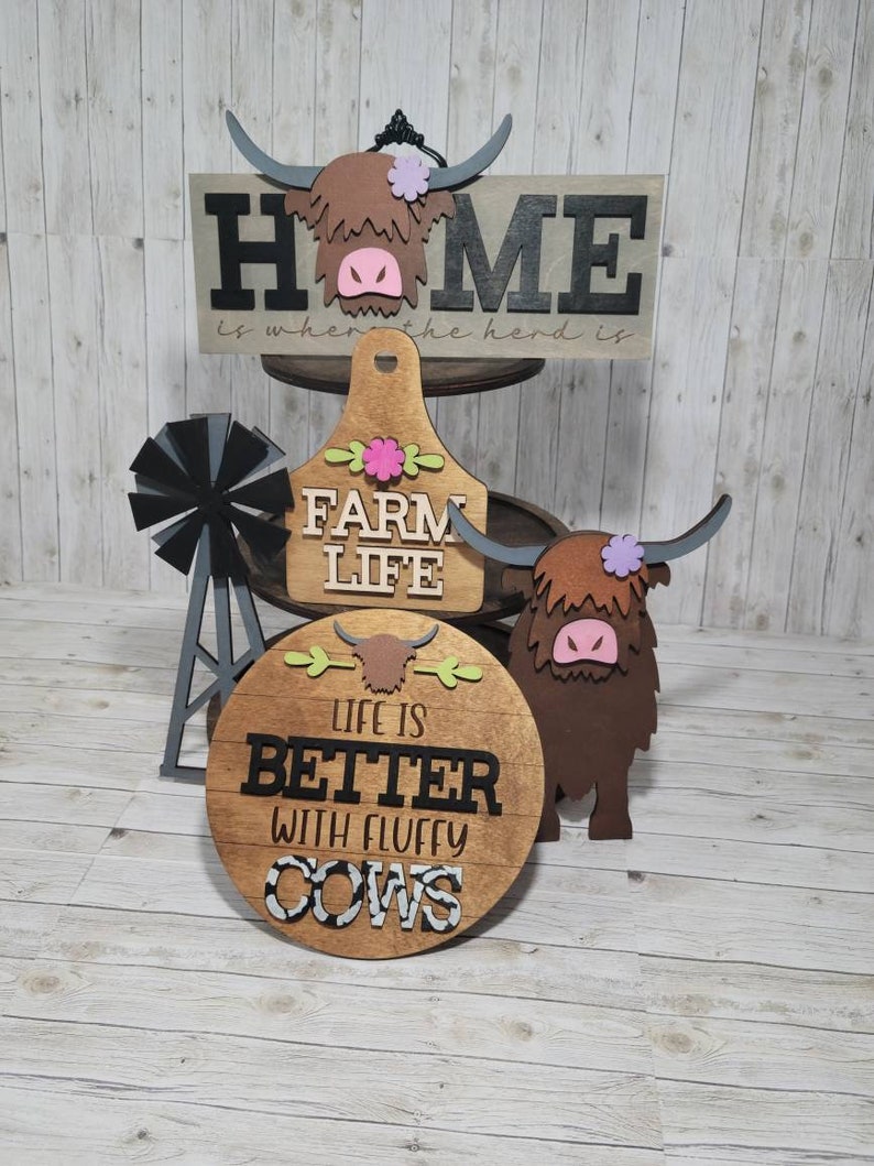 Op de afbeelding: Een set van drie houten borden met een boerderijthema. Het bovenste bord staat "Home is where the herd is". Het middelste bord staat "Farm Life" en het onderste bord staat "Life is better with fluffy cows".