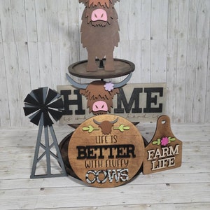 Op de afbeelding: Houten decorset voor thuis met een bruine hooglandkoe, een bord met de tekst "Home is where the herd is", een rond bord met de tekst "Life is better with fluffy cows" en een houten label met de tekst "Farm Life".