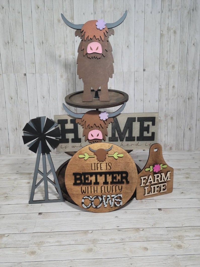 Op de afbeelding: Houten decorset voor thuis met een bruine hooglandkoe, een bord met de tekst "Home is where the herd is", een rond bord met de tekst "Life is better with fluffy cows" en een houten label met de tekst "Farm Life".