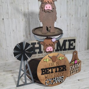 Op de afbeelding: Een drielaags houten taartstandaard met een boerderijthema. De bovenste laag heeft een bruine hooglandkoe met een roze neus en een paarse bloem. De middelste laag heeft een houten bord met de tekst "Home is where the herd is". De onderste laag heeft een rond houten bord met de tekst "Life is better with fluffy cows" en een kleiner houten bord met de tekst "Farm Life".