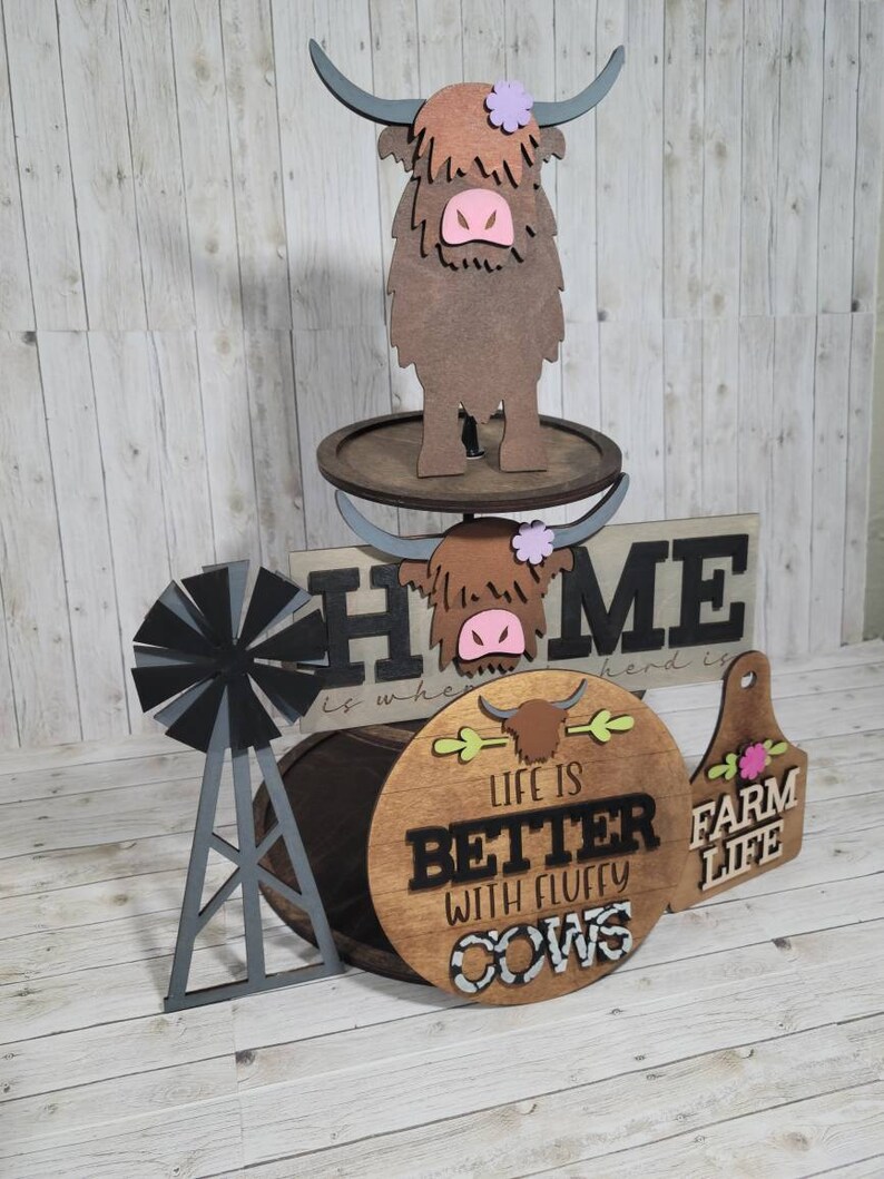 Op de afbeelding: Een drielaags houten taartstandaard met een boerderijthema. De bovenste laag heeft een bruine hooglandkoe met een roze neus en een paarse bloem. De middelste laag heeft een houten bord met de tekst "Home is where the herd is". De onderste laag heeft een rond houten bord met de tekst "Life is better with fluffy cows" en een kleiner houten bord met de tekst "Farm Life".