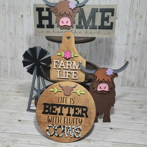 Op de afbeelding: Houten boerderijdecor met een hooglandkoe-thema. De borden lezen "Home is where the herd is", "Farm Life" en "Life is better with fluffy cows".