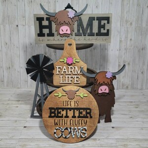 Op de afbeelding: Drie houten borden met een boerderijthema. Het bovenste bord zegt "Home is where the herd is" met een bruine Highland-koeienkop-uitsparing. Het middelste bord zegt "Farm Life" met een roze bloemafbeelding. Het onderste bord zegt "Life is better with fluffy cows" met een zwart-witte koeienprint.
