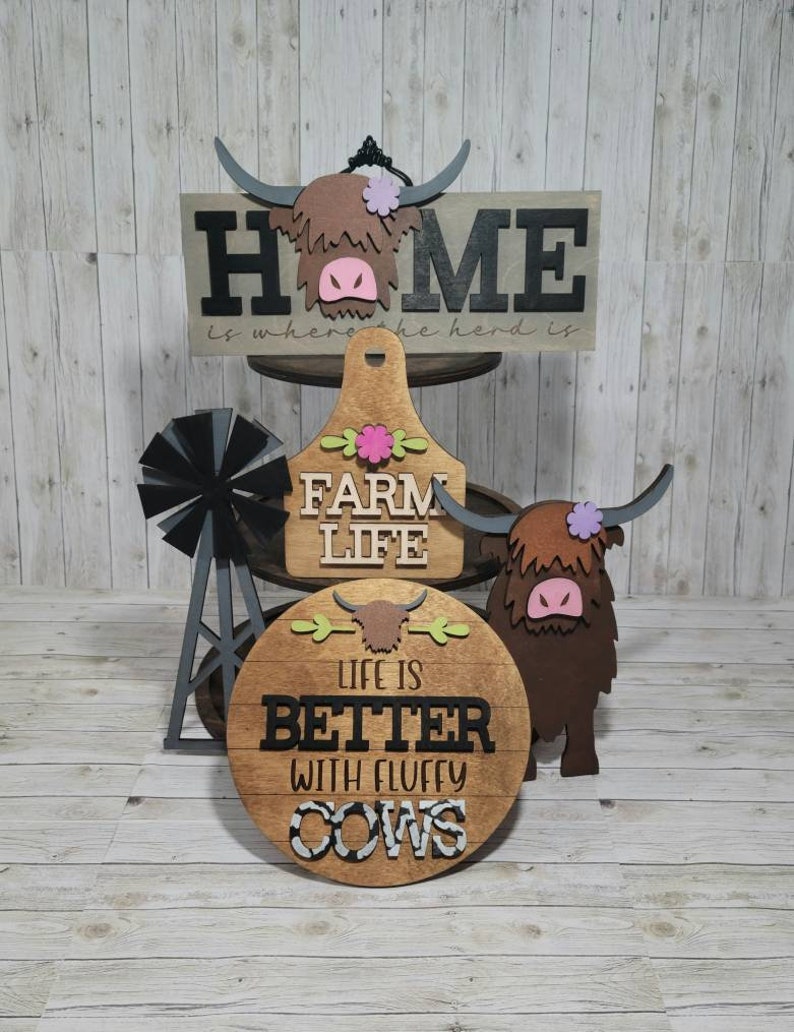 Op de afbeelding: Drie houten borden met een boerderijthema. Het bovenste bord zegt "Home is where the herd is" met een bruine Highland-koeienkop-uitsparing. Het middelste bord zegt "Farm Life" met een roze bloemafbeelding. Het onderste bord zegt "Life is better with fluffy cows" met een zwart-witte koeienprint.