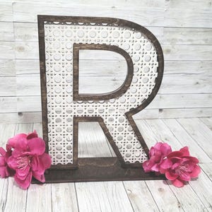 May include: A wooden letter "R" with a dark brown frame and a white patterned interior. The letter is standing upright, with pink flowers at the base. The background is a light wood grain.