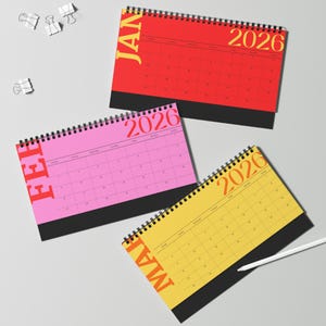 May include: Three colorful 2026 desk calendars with spiral binding. The calendars are red, pink, and yellow, each displaying a different month. The word "JAN" is printed vertically on the red calendar, "FEB" on the pink, and "MAR" on the yellow.