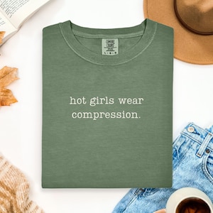 May include: Sage green t-shirt with the text "hot girls wear compression." The shirt is a crew neck and appears to be made of a soft material. A brown hat, open book, and blue jeans are in the background.